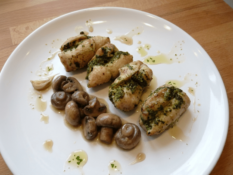 Roladki z kurczaka z pesto ułożone na białym talerzu skropionym sosem, podane z duszonymi pieczarkami.